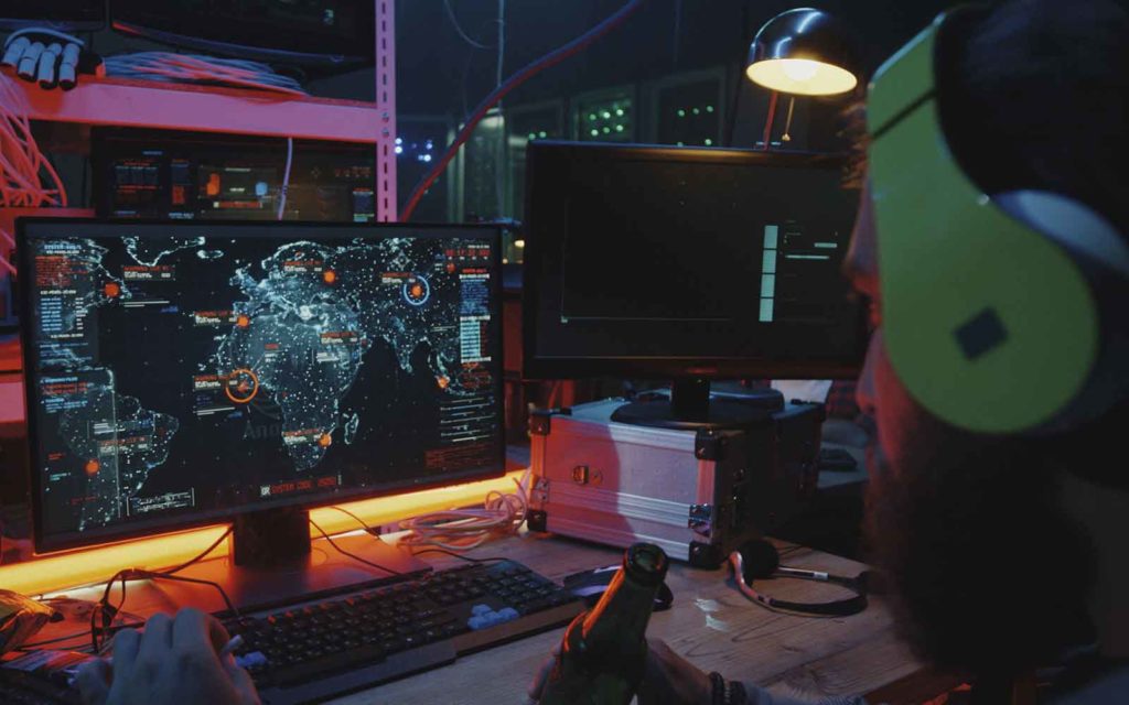 hacker motivation graphic is a photograph of a hacker in front of a computer with locations he's targeting with cyber attacks on a map